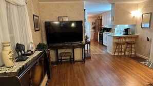 A living room with a flat-screen TV, wooden flooring, a dining area with a table and chairs, and a kitchen with bar stools.