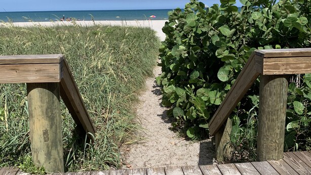 Een houten pad dat naar een strand leidt, met een helder uitzicht op de oceaan en een zandstrand in de verte.
