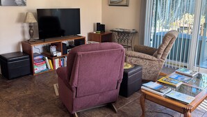 Een woonkamer met een televisie, boeken, een paarse fauteuil, een glazen salontafel en een plafondventilator.