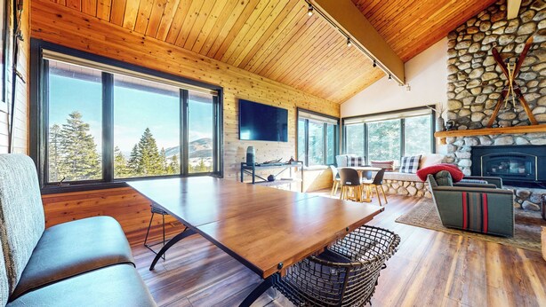 A spacious living area with a large wooden table, comfortable seating, a stone fireplace, and large windows offering a view of trees and mountains. - Remodeled Home, Amazing Views, Private Hot Tub & Garage, Near the Lakes Basin (Mammoth Lakes)