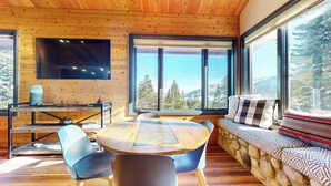 A cozy cabin interior with a wooden ceiling, a large TV mounted on the wall, a round dining table with chairs, a stone fireplace, and a sofa with a view of the forest and mountains through large windows. - Remodeled Home, Amazing Views, Private Hot Tub & Garage, Near the Lakes Basin (Mammoth Lakes)