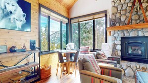 A cozy cabin interior with a stone fireplace, wooden walls, a dining area, and a large window offering a view of trees. - Remodeled Home, Amazing Views, Private Hot Tub & Garage, Near the Lakes Basin (Mammoth Lakes)