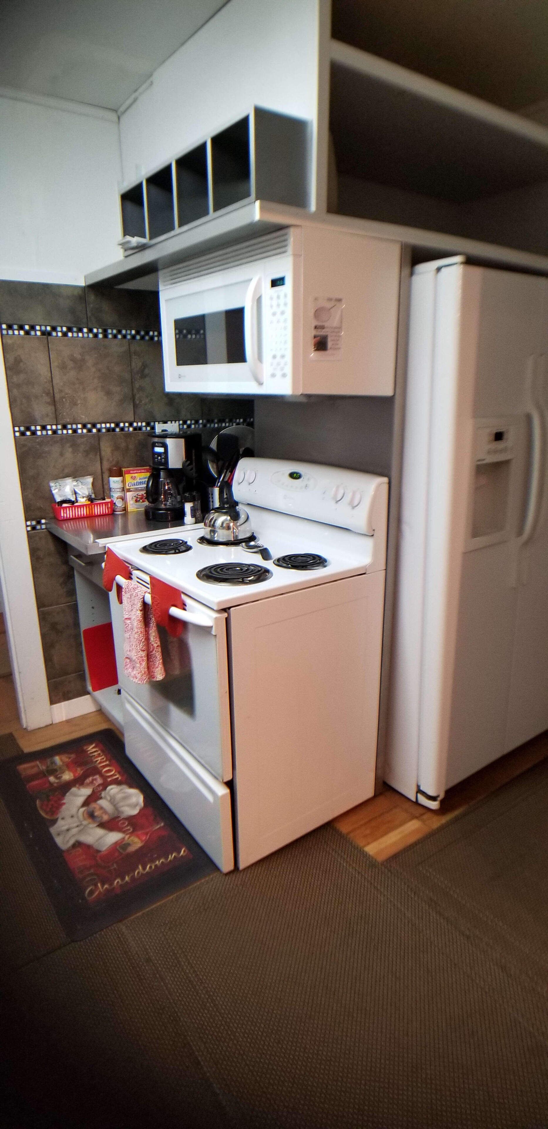 Une cuisine compacte avec un four, un micro-ondes et un réfrigérateur blancs. Une serviette à carreaux rouges et blancs est accrochée à la poignée du four. Une machine à café et une boîte de capsules de café sont posées sur le plan de travail.
