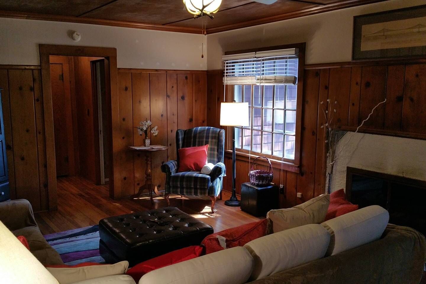A cozy living room with wooden paneling, a plaid armchair, a black ottoman, and a fireplace.