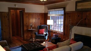 Una sala de estar acogedora con paneles de madera, un sillón de cuadros, un reposapiés negro y una chimenea.
