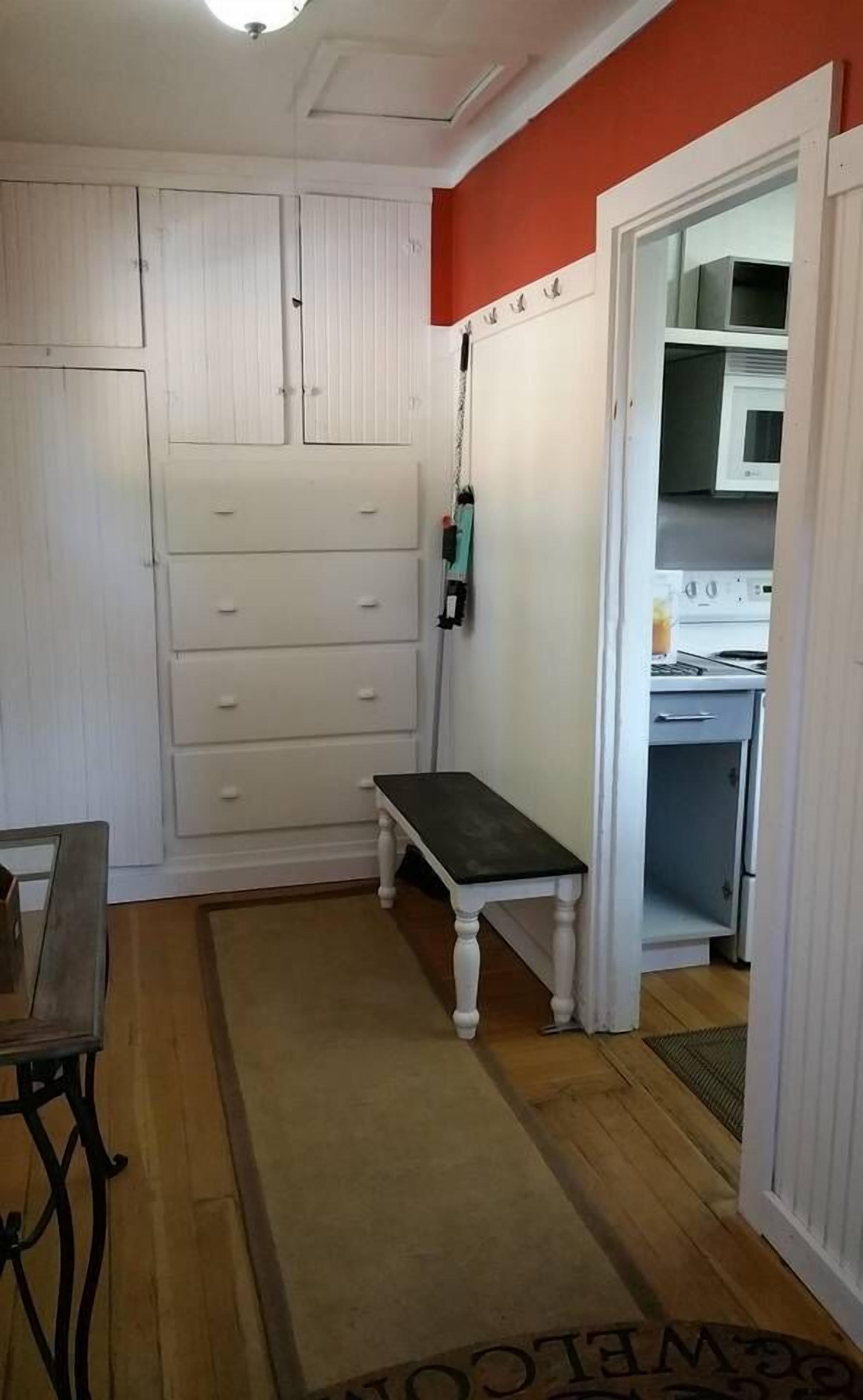 A room with a white wardrobe, a bench, and a kitchen visible through an open door.