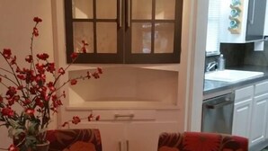 Un comedor con una mesa redonda, sillas estampadas con motivos florales rojos, un jarrón con flores rojas y un aparador con puertas de vidrio.