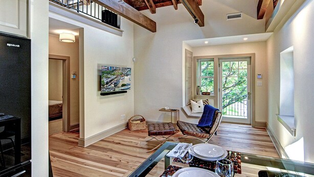 Une pièce spacieuse avec un plafond haut, des poutres en bois, un coin repas avec une table dressée pour deux personnes, un fauteuil confortable et une vue sur l’extérieur grâce à une porte vitrée.