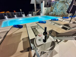 A poolside area with lounge chairs, a table with a bottle and glass, and a view of the sea at night. - Ivan's holiday house with private swimming pool (Klis)