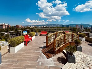 A rooftop terrace with a wooden bridge, red benches, potted plants, and a cityscape view. - Suites Terraza Imperial (Guatemala City)