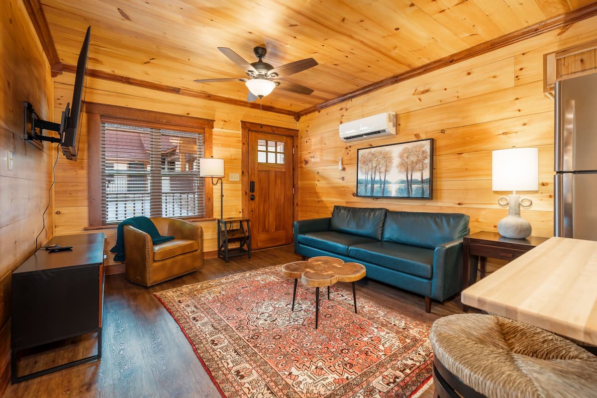 A cozy log cabin interior with a wooden ceiling, a ceiling fan, a blue sofa, a small coffee table, a leather armchair, a wooden side table, and a rug.