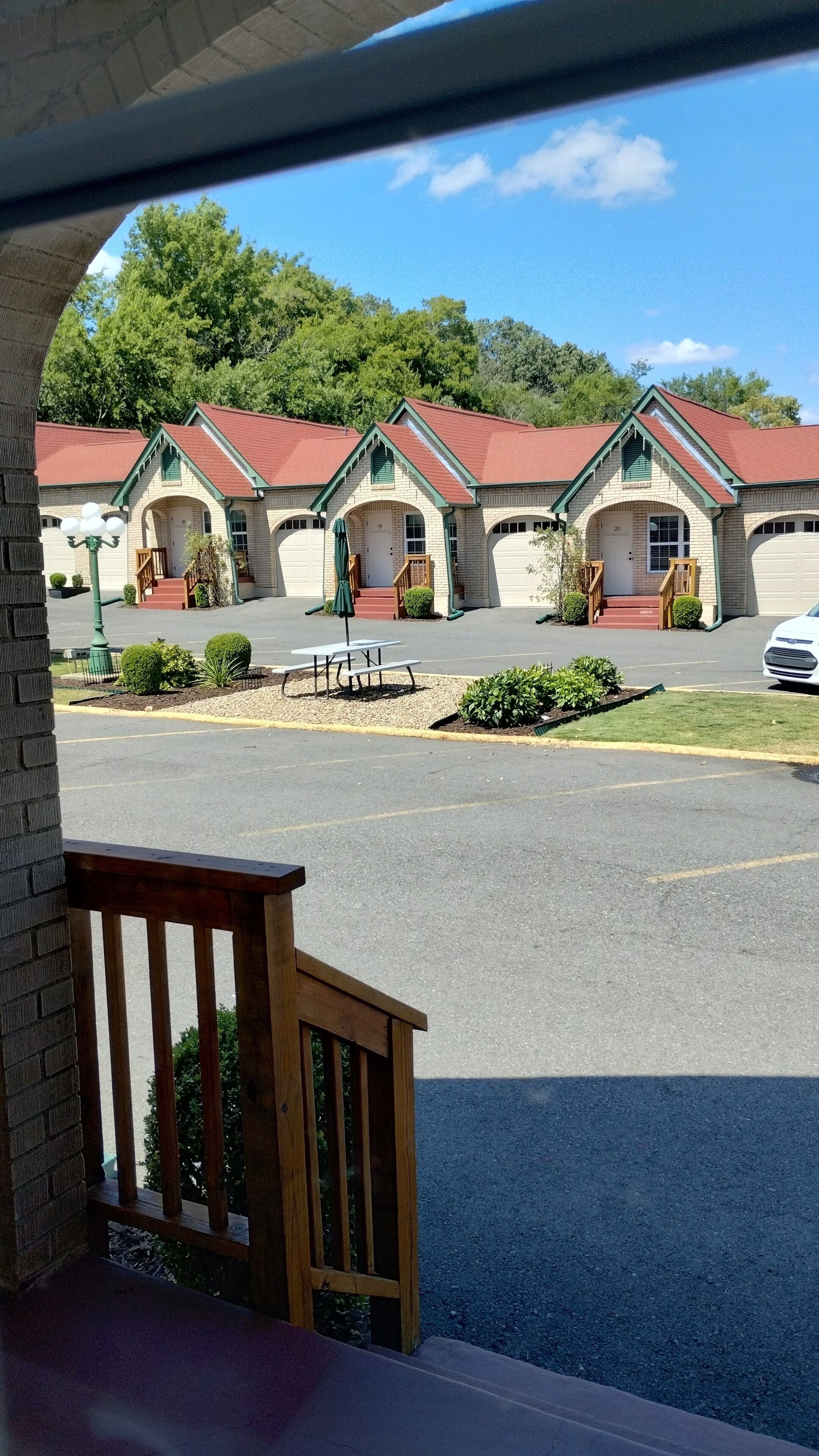 A motel with multiple buildings, each featuring a red roof and green trim. There is a parking lot in front with a picnic table and benches.