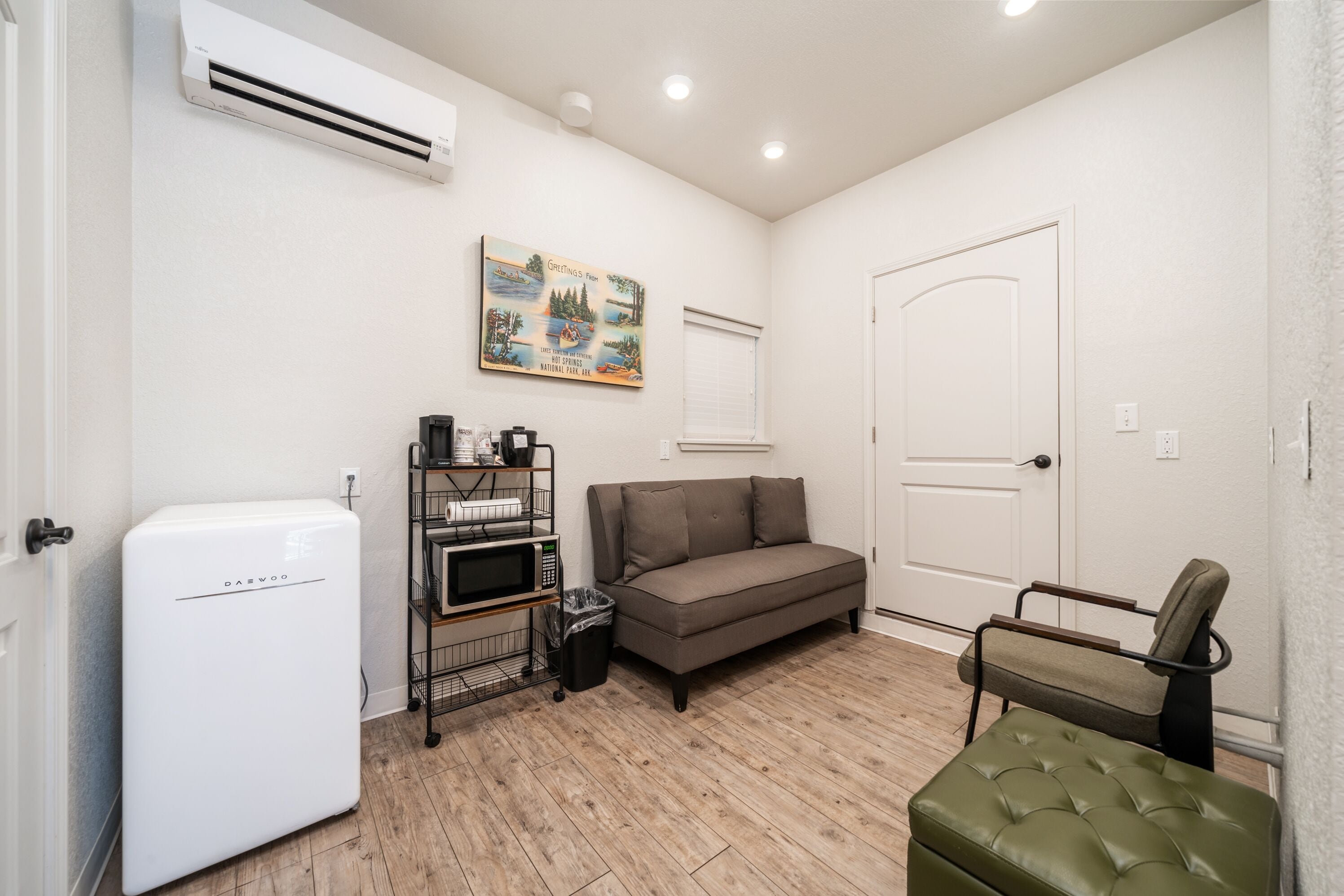 Um espaço compacto com ar condicionado branco, sofá, um banquinho verde, micro-ondas e uma lixeira pequena.