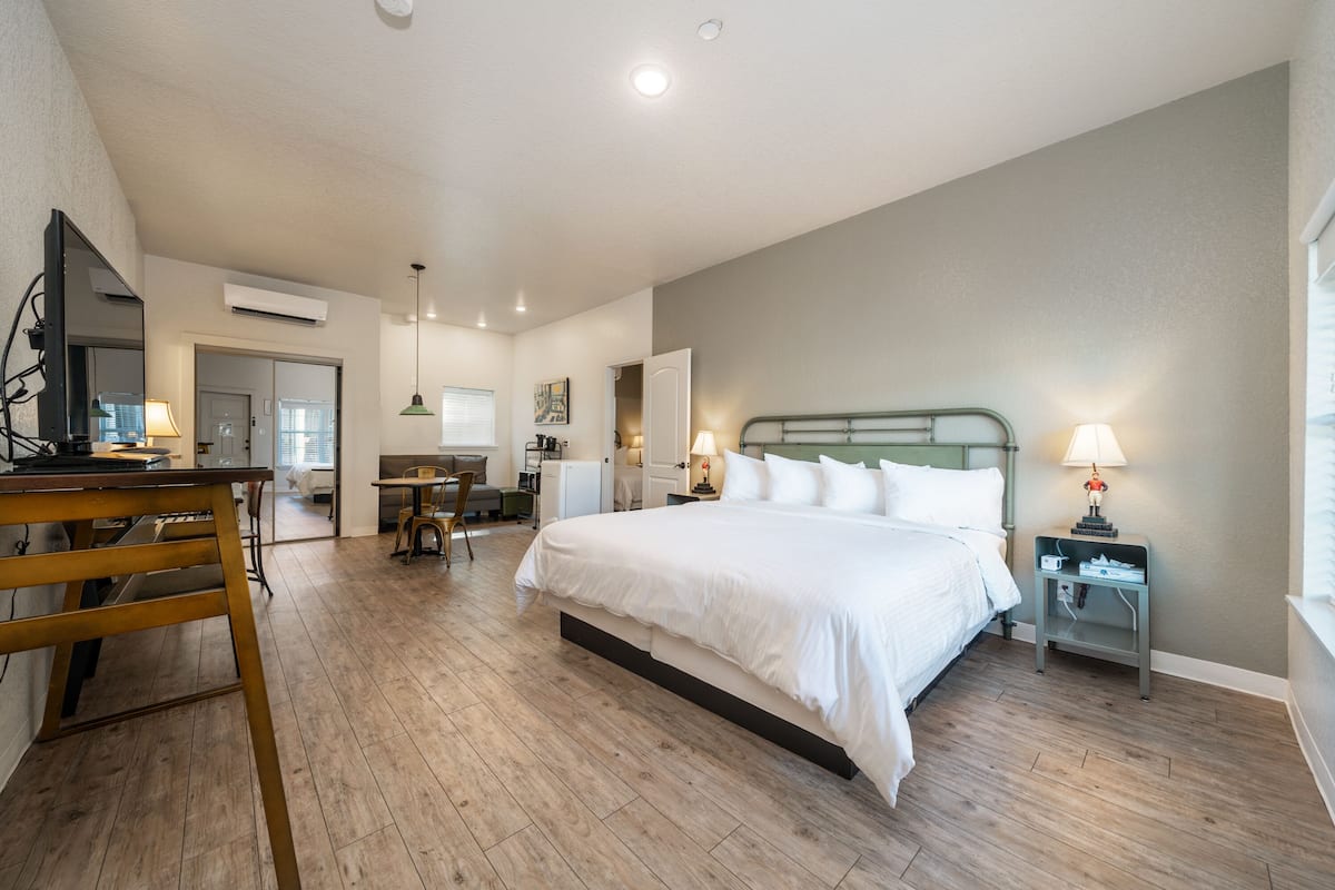 A hotel room with a large bed, a desk, a chair, a small table, and a nightstand with a lamp.