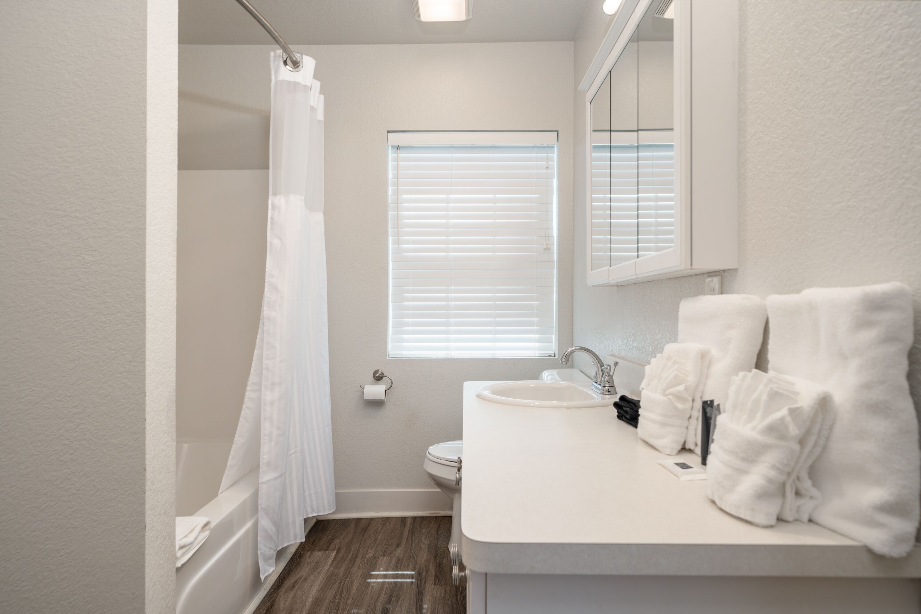 A bathroom with a shower, toilet, sink, and a window with blinds.