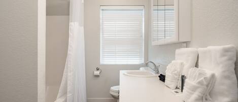 A bathroom with a shower, toilet, sink, and a window with blinds.