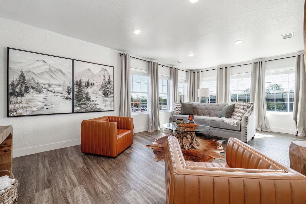 A spacious living room with a grey sofa, orange armchair, and a coffee table with a wooden centerpiece. Large windows offer a view of the outdoors.