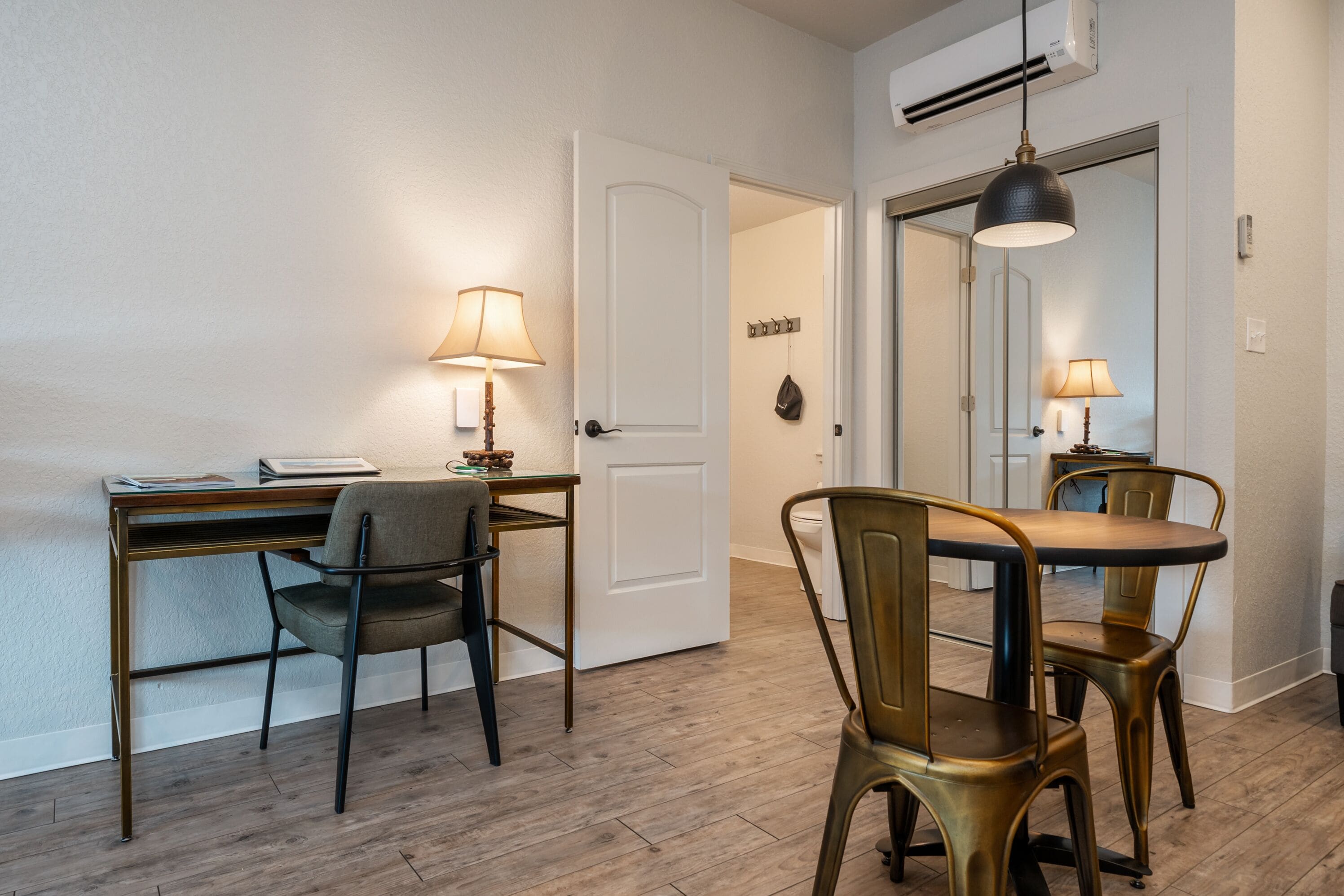 A room with a wooden desk, two chairs, a dining table with chairs, a lamp, and a door.