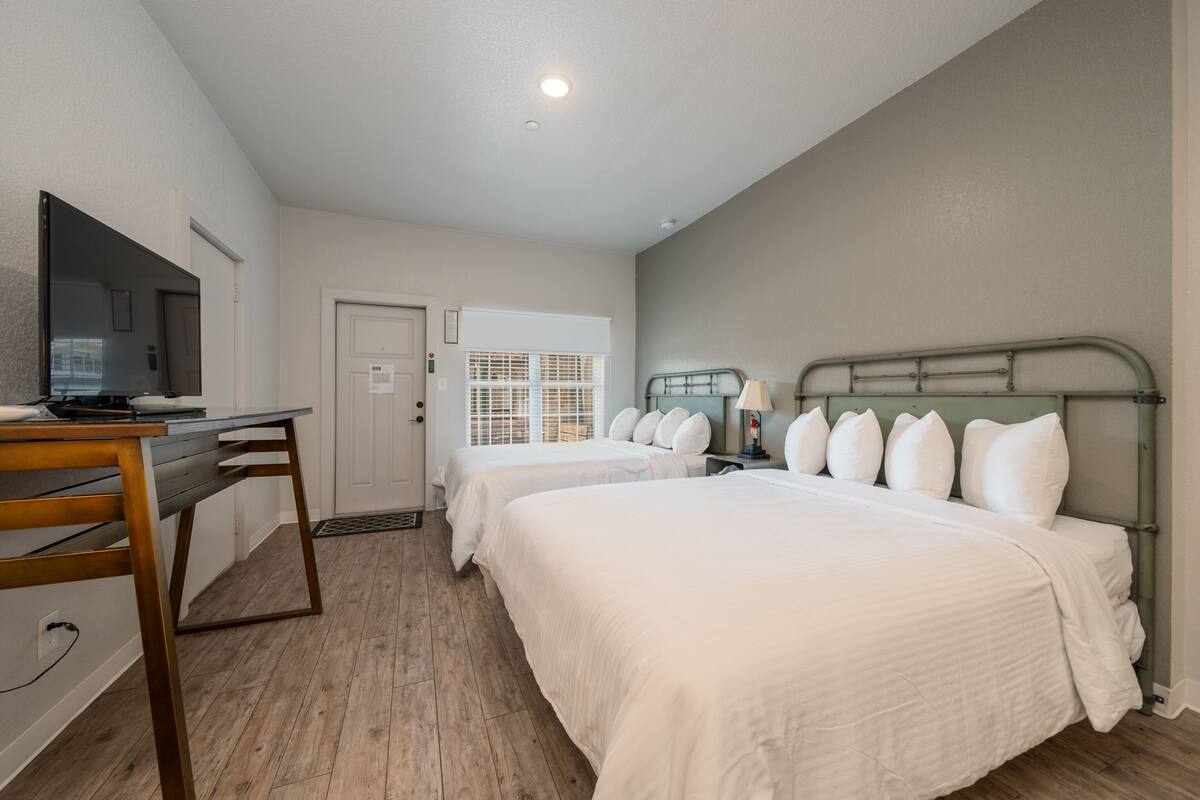 A hotel room with two single beds, a desk with a chair, a television, and a window with blinds.