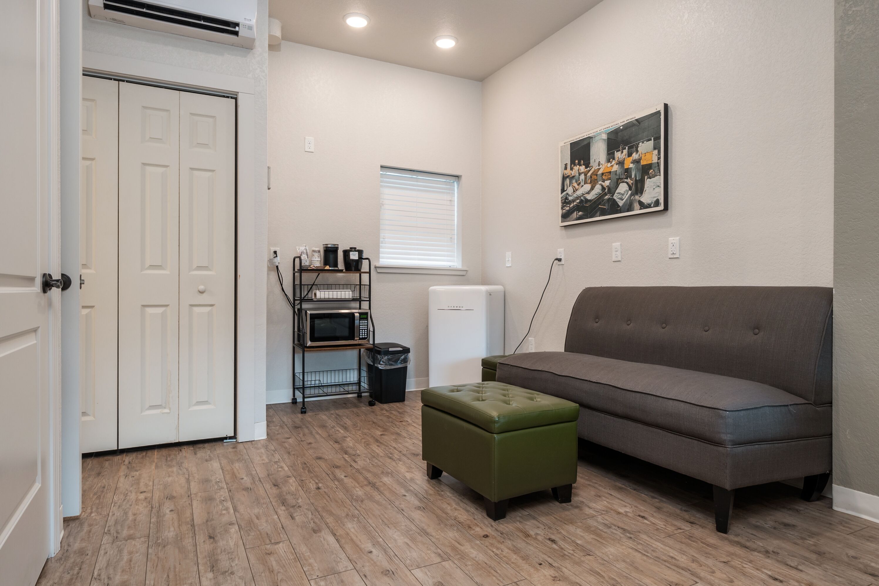 Una habitación con sofá gris, reposapiés verde, microondas, refrigerador, ventana con persianas y puerta con armario blanco.