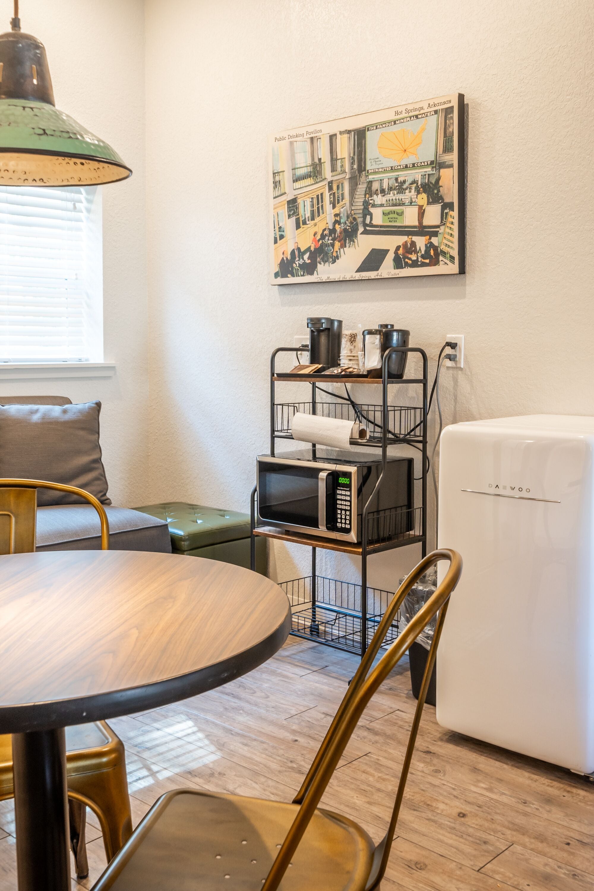 Una kitchenette compacta con microondas, un refrigerador y una repisa metálica con ollas y sartenes. Hay una mesa de comedor con sillas, un sofá y una lámpara verde. Un cuadro enmarcado con una escena de pueblo cuelga en la pared.