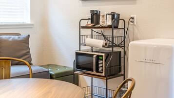 Una kitchenette compacta con microondas, un refrigerador y una repisa metálica con ollas y sartenes. Hay una mesa de comedor con sillas, un sofá y una lámpara verde. Un cuadro enmarcado con una escena de pueblo cuelga en la pared.