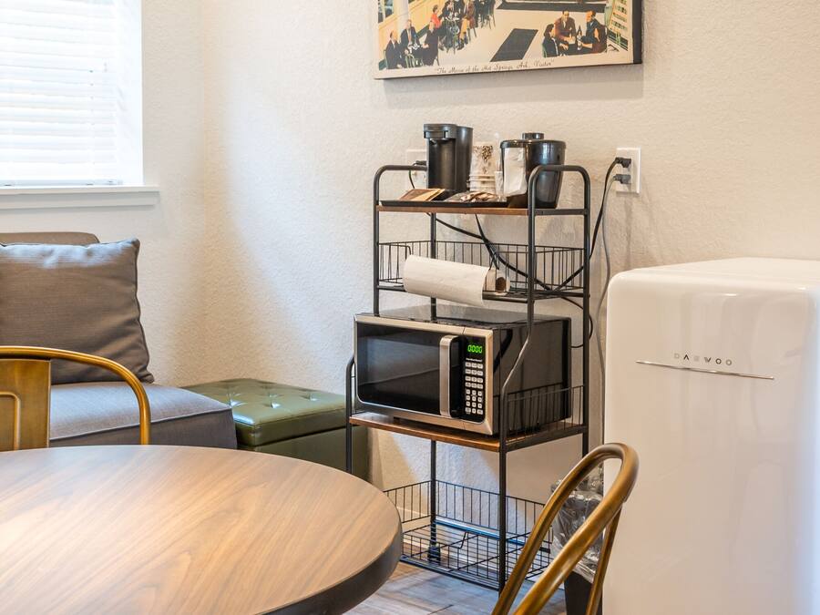 Una kitchenette compacta con microondas, un refrigerador y una repisa metálica con ollas y sartenes. Hay una mesa de comedor con sillas, un sofá y una lámpara verde. Un cuadro enmarcado con una escena de pueblo cuelga en la pared.