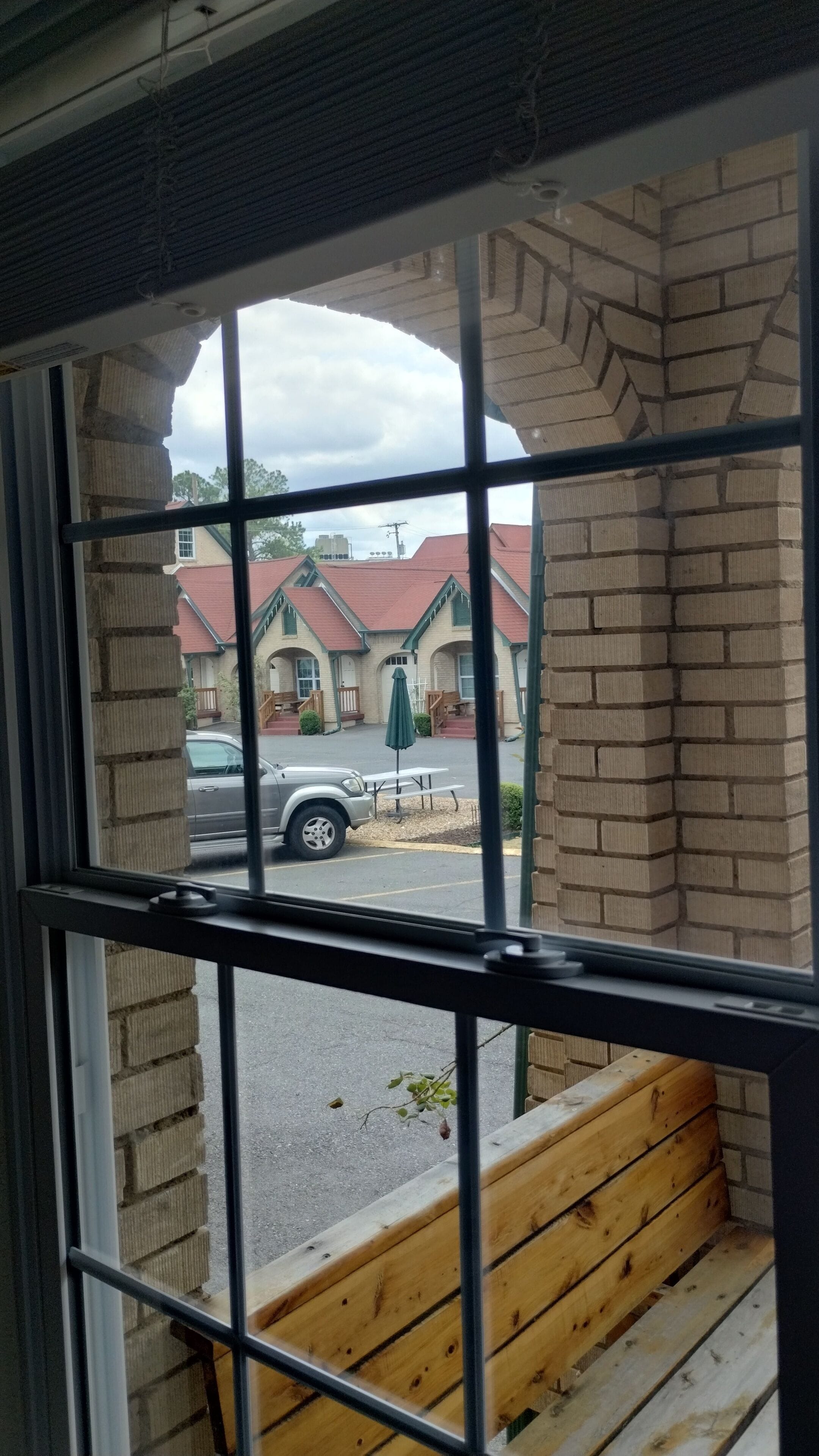 A view through a window with a metal grid, featuring a brick building with a red roof, a parked car, and a wooden bench outside.