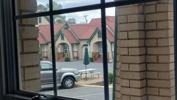 Visão pela janela com grade de metal, mostrando um edifício de tijolos com telhado vermelho, um carro estacionado e um banco de madeira na parte externa.