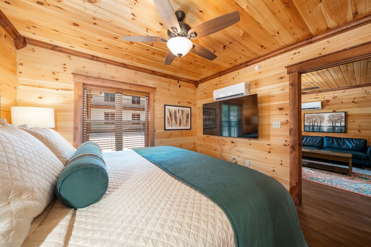 A wooden cabin bedroom with a bed, a ceiling fan, a TV, and a window with blinds.