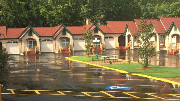 A row of motel buildings with red roofs and green trim, a parking lot with yellow lines, and a small green area with a bench. - Best  Court (Hot Springs)