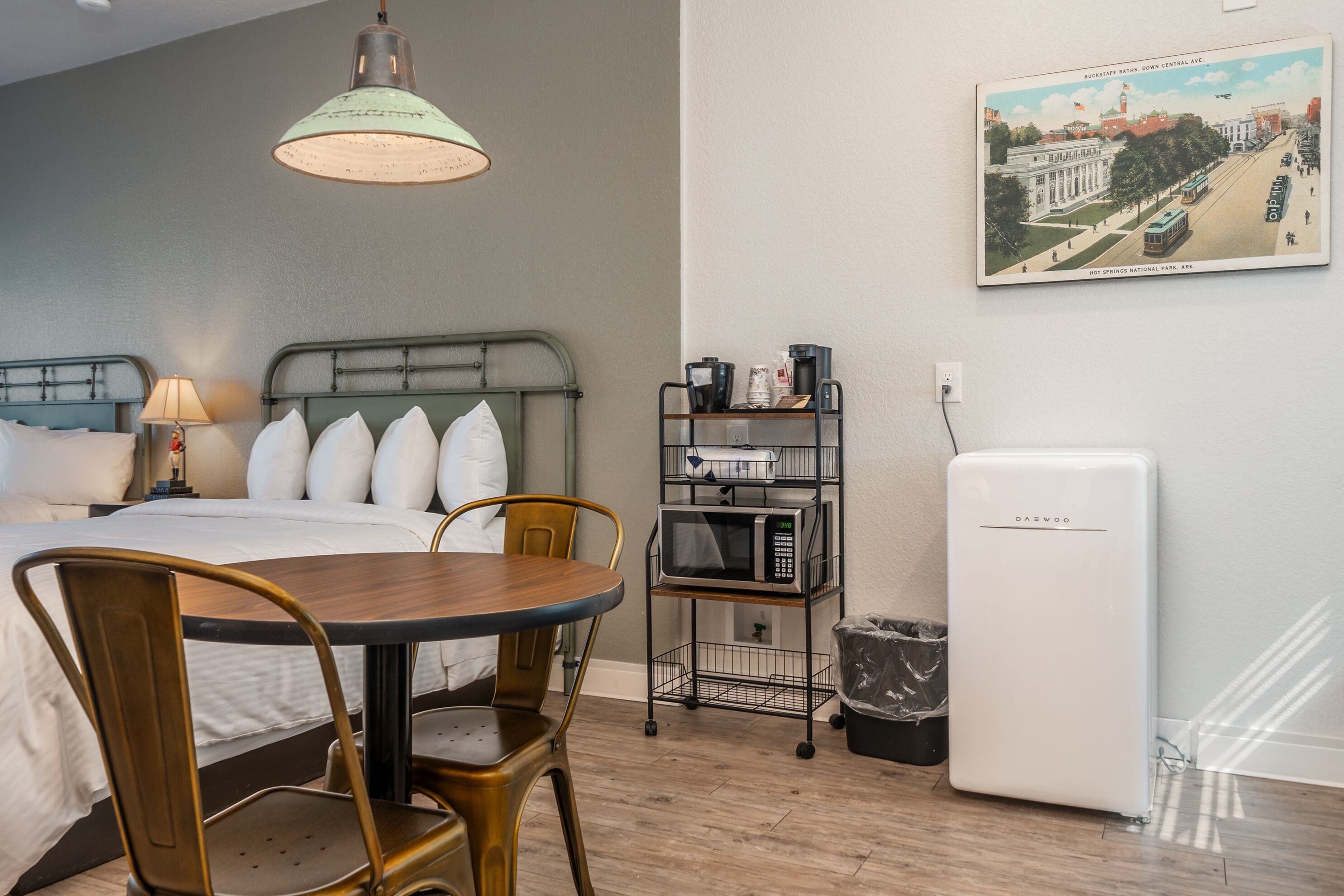 A hotel room with a bed, a dining table with chairs, a microwave, a refrigerator, a trash bin, a lamp, and a wall-mounted picture of a cityscape.