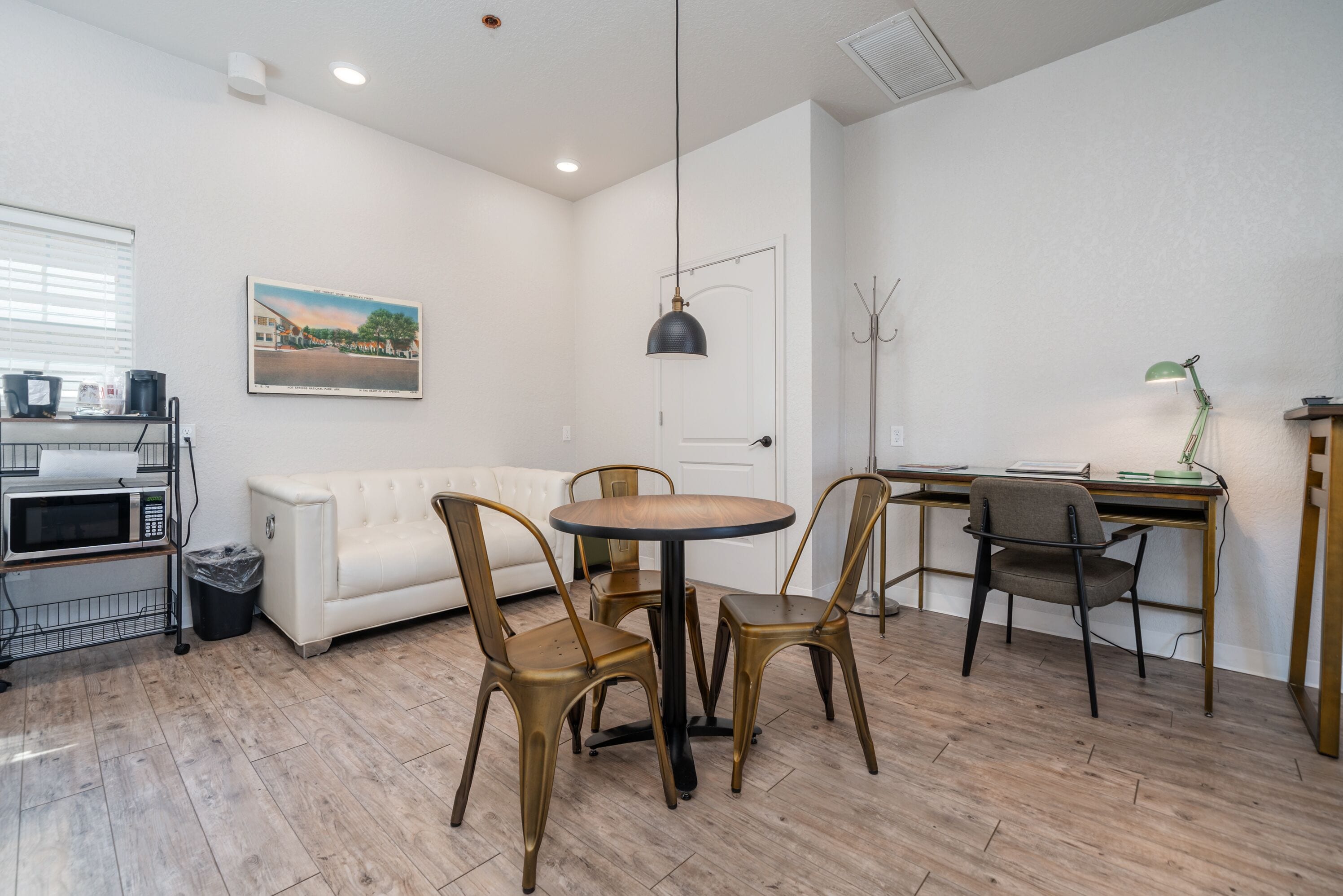 Un espacio habitable pequeño con una mesa redonda de madera, dos sillas, un sofá blanco, un escritorio con una computadora y un cesto de basura.
