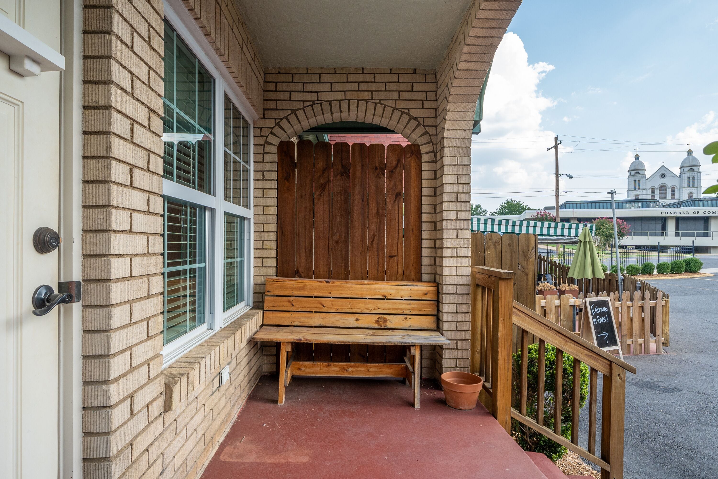 A wooden bench outside a building with a brick facade, a wooden fence, and a sign pointing towards the entrance.