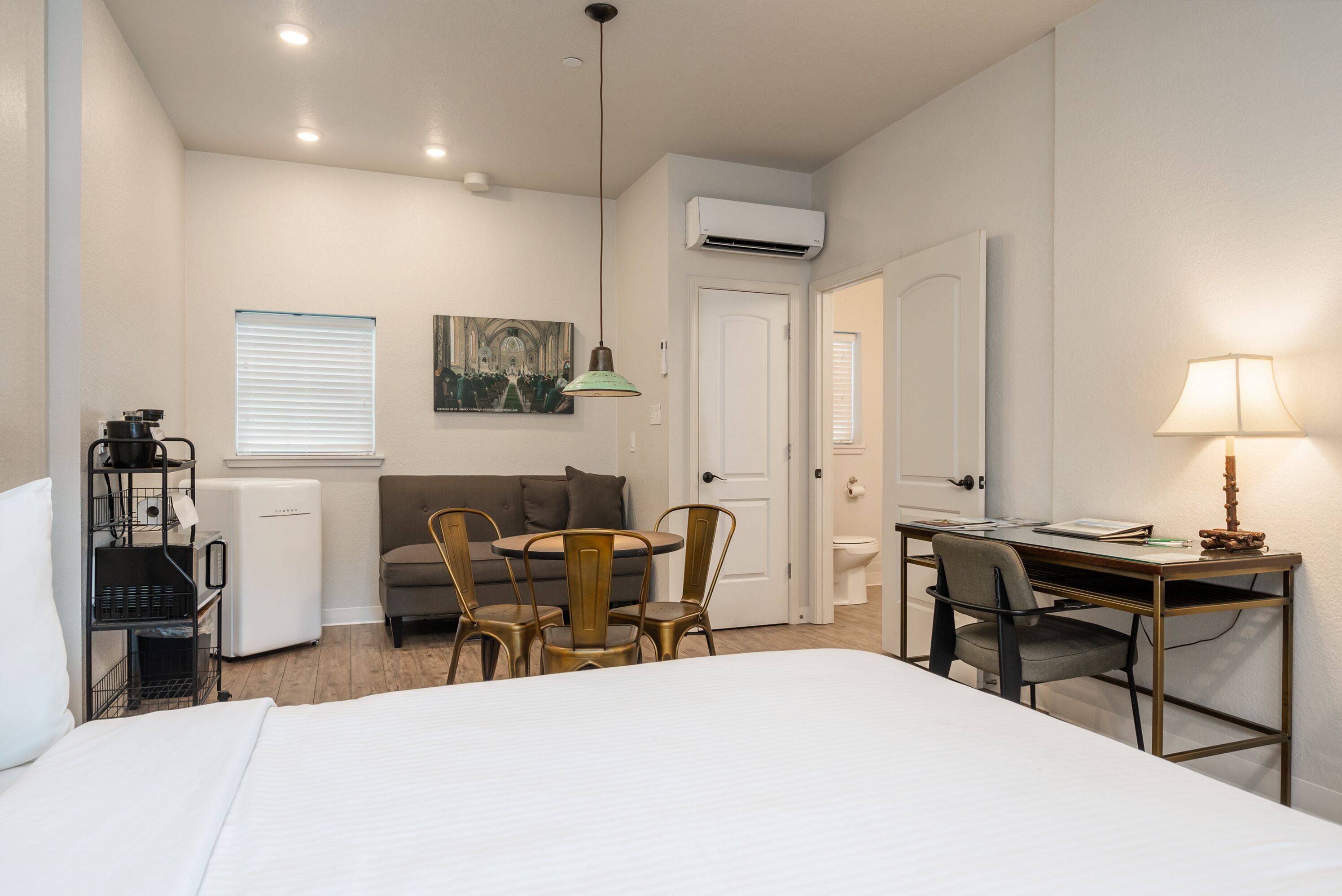 Une chambre d’hôtel comprenant un lit, un bureau avec une chaise, une petite table à manger avec des chaises, un coin cuisine et un tableau au mur.