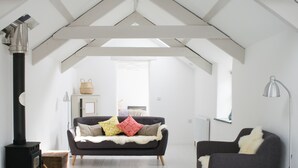 Ett modernt sovrum i loftstil med högt i tak, synliga takbjälkar, en central soffa med mönstrade kuddar, en fåtölj, en golvlampa och en vedkamin.