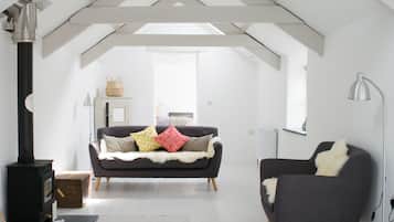 Un dormitorio estilo loft moderno con techo alto, vigas a la vista, un sofá central con cojines estampados, una butaca, una lámpara de pie y un estufa de leña.