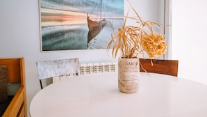 Un coin repas avec une table blanche, un vase contenant des plantes séchées, une chaise en bois et un tableau représentant un bateau sur un quai.