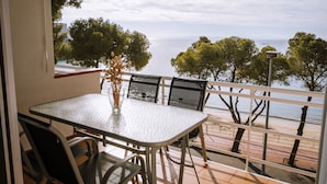 Un balcon avec une table et des chaises, donnant sur une plage bordée d’arbres et offrant une vue sur un ciel dégagé.