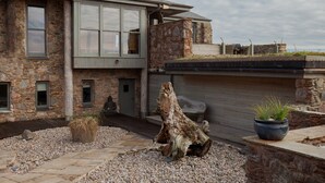 Een modern stenen huis met een groot raam, een beeld en een ruwe boomstronk in de voortuin.