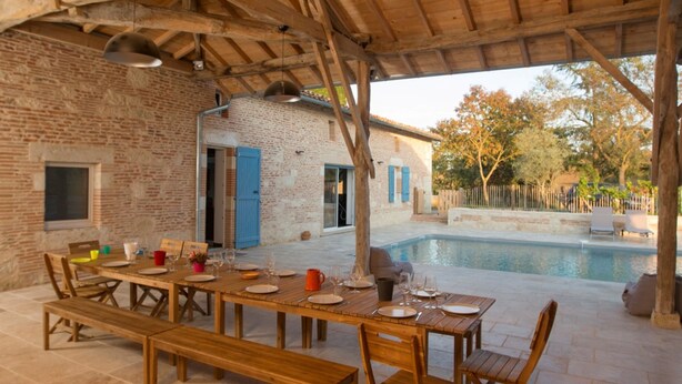 An outdoor dining area with a wooden table and chairs, a brick wall, and a pool in the background. - AN AIR OF DETENTE **** (4 ears and 4 stars) (Faudoas)