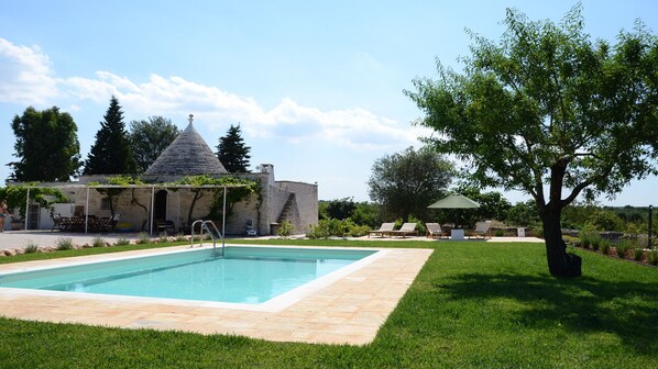 Una piscina con bordo in pietra, un portico coperto con posti a sedere e un edificio con tetto a cono.