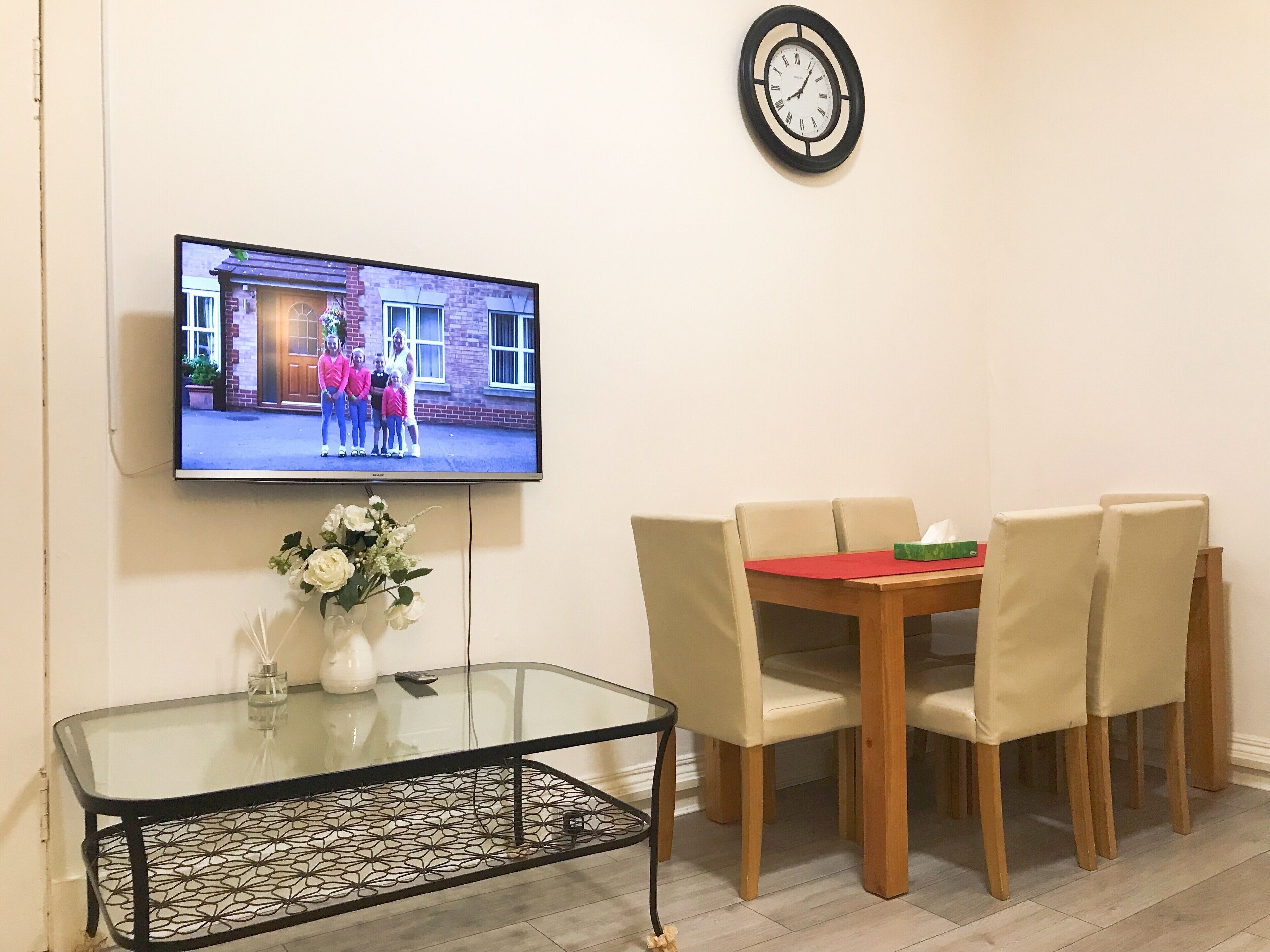 A dining area with a glass-top table, beige chairs, a clock on the wall, and a TV displaying a family photo.