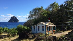 Um pequeno edifício térreo com telhado simples e uma torre decorativa, rodeado por vegetação natural e rochas, com vista para o oceano e uma ilha distante.