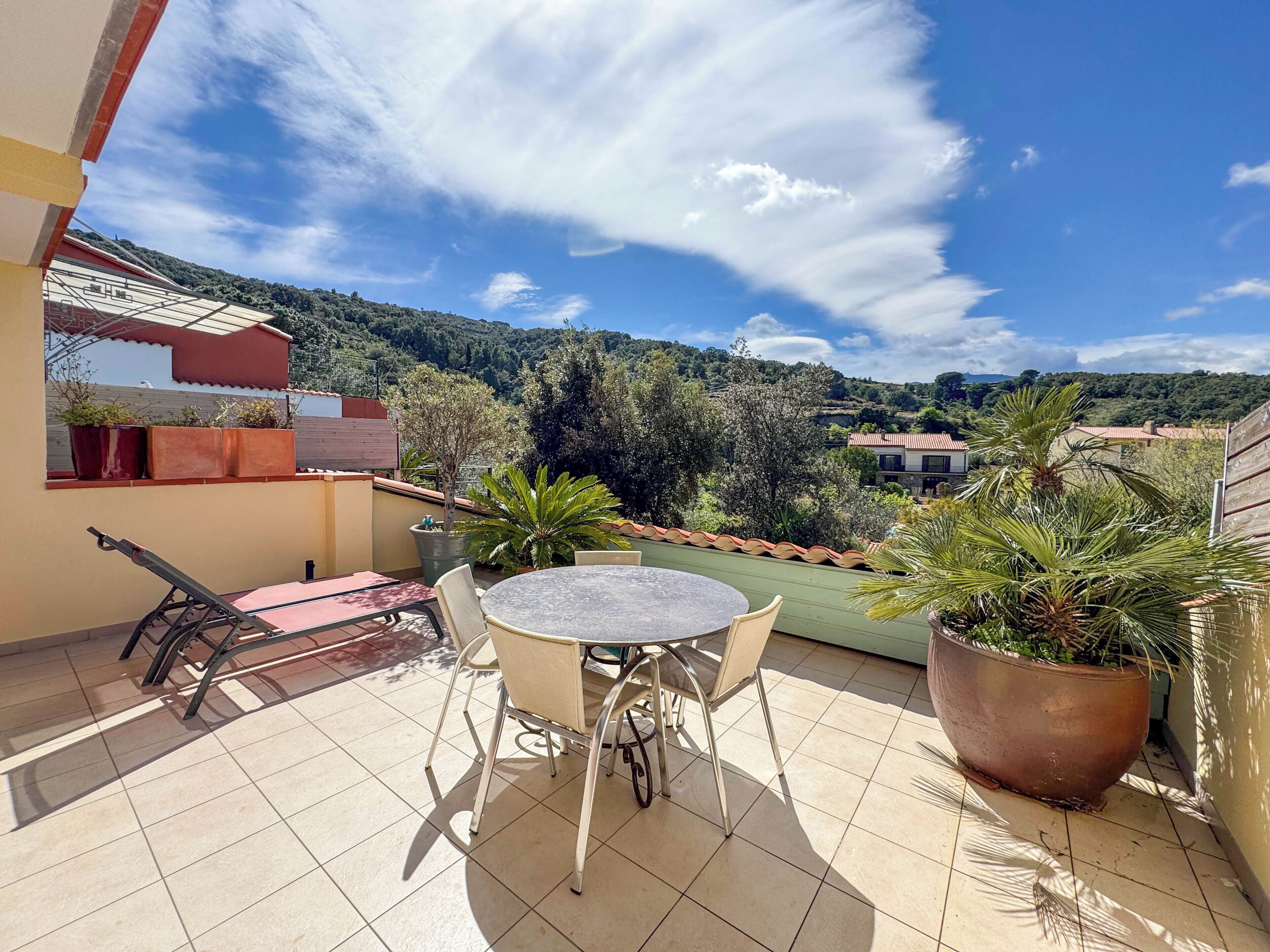 Un balcon avec une table ronde et des chaises, des plantes en pot et une vue sur un paysage vallonné.