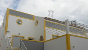 A building with a staircase leading to a landing, a covered outdoor area, and a flat roof with antennas. - New 3-bedroom apartment 50 meters from the beach (Vila Real de Santo António)