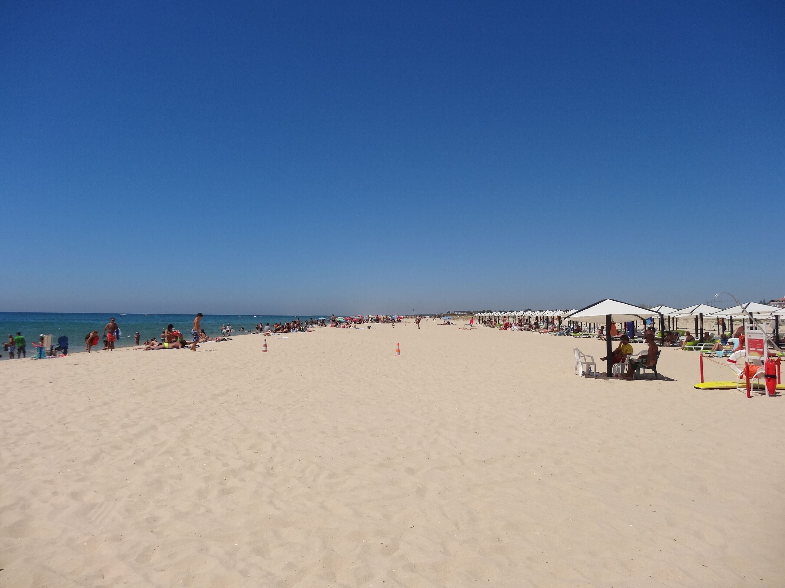 Une vaste plage de sable, parsemée de parasols et de chaises longues, un ciel bleu azur et l’océan en toile de fond.