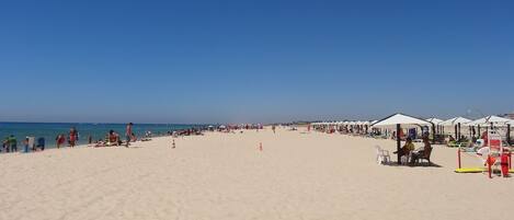 Une vaste plage de sable, parsemée de parasols et de chaises longues, un ciel bleu azur et l’océan en toile de fond.