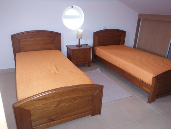 Quarto com duas camas de madeira, uma mesinha de cabeceira com abajur e um espelho redondo na parede.