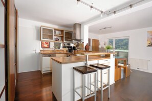 Une cuisine moderne avec des plans de travail en bois, un îlot central, des tabourets de bar et une vue sur un lac, visible à travers une grande fenêtre.